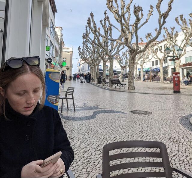 Rosemary Richings in a black jacket with sunglasses on her head. She's looking at her smartphone on a black chair with another chair beside her. Branchless trees are in the background on cobblestone streets. Crowds of people are walking behind her in all directions. A grey brick building is behind her.